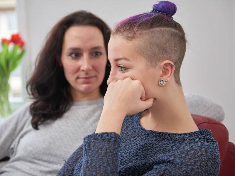 Zwei Personen sitzen auf einem Sofa, wobei die im Vordergrund nachdenklich wirkt und die andere sie ruhig ansieht, im Hintergrund sind rote Blumen auf einem Tisch zu sehen.
