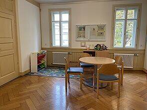 Ein helles Zimmer mit Holzboden, einem runden Tisch und Stühlen, Spielzeug in der Ecke und Fenstern mit Blick auf Bäume.