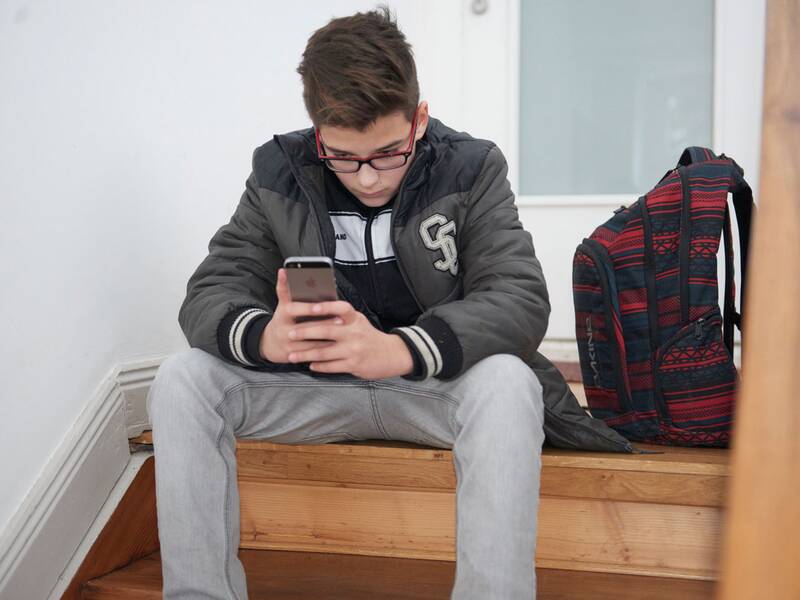 Eine Person mit Brille sitzt auf einer Treppe und schaut auf ein Smartphone, während neben ihr ein bunter Rucksack steht.
