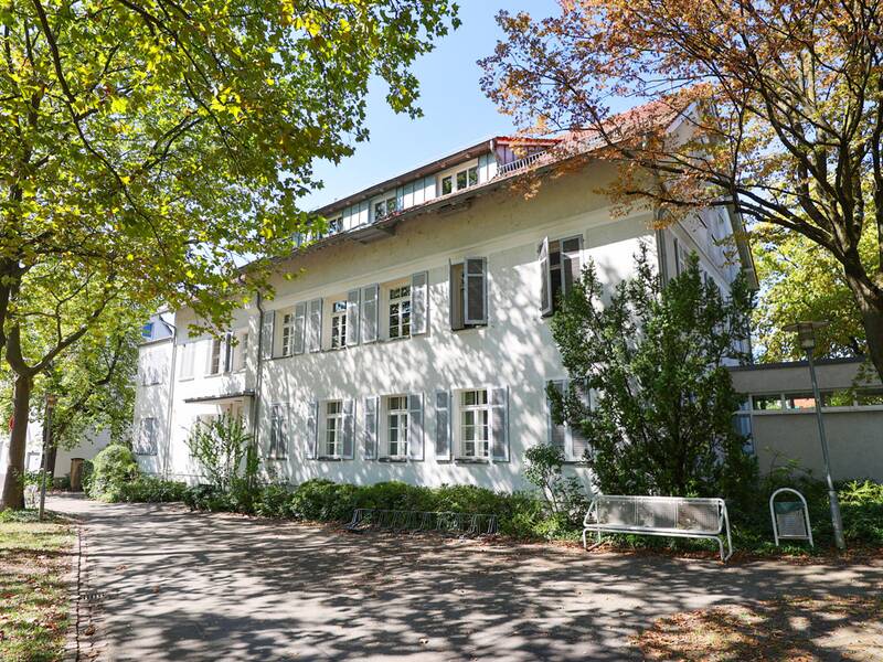 Musikschule. Ein zweistöckiges, weißes Gebäude mit Sprossenfenstern steht von Bäumen umgeben an einem sonnigen Tag.