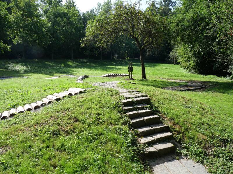 Eine kleine Steintreppe führt über eine Wiese zu einem Baum, dahinter verlaufen verschiedene Barfußpfad-Elemente.