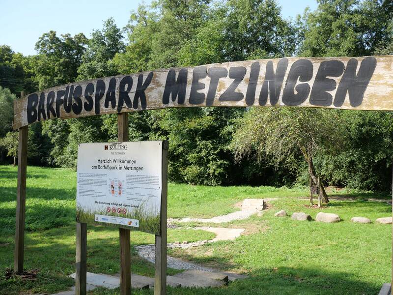 Ein hölzernes Eingangsschild mit der Aufschrift „Barfußpark Metzingen“ steht über einer Infotafel vor grüner Wiese und Bäumen.