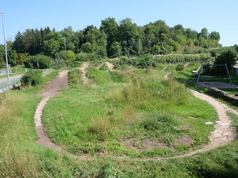 Dirtpark mit gewundenen Wegen, umgeben von Bäumen und einem Spielplatz im Hintergrund.