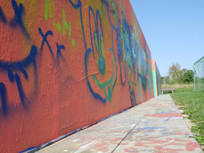 Eine orangefarbene Wand mit einfachen, bunten Graffiti-Zeichnungen, darunter Comicgesichter und bunte Linien. Der Boden ist ebenfalls mit Farbflecken und Mustern übersät, im Hintergrund Wiese und blauer Himmel.