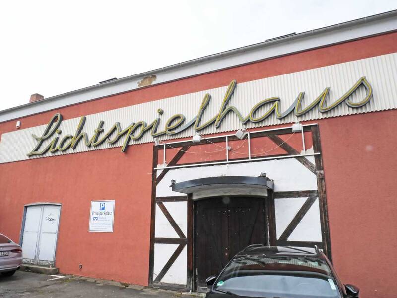 Das Bild zeigt die Fassade eines alten Gebäudes mit der Aufschrift "Lichtspielhaus" über einem dunklen Holztor, vor dem ein Auto parkt.