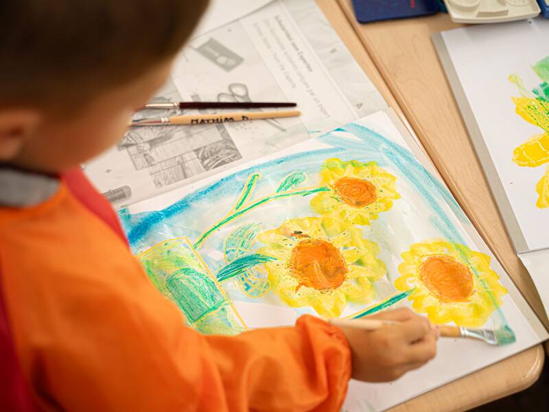 Ein Kind malt konzentriert mit Pinsel und leuchtenden Farben Sonnenblumen auf ein Blatt Papier.