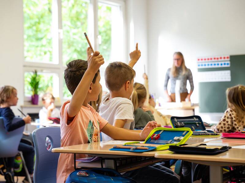 Kinder sitzen in einem Klassenzimmer und heben ihre Hände, während die Lehrerin vorne steht.