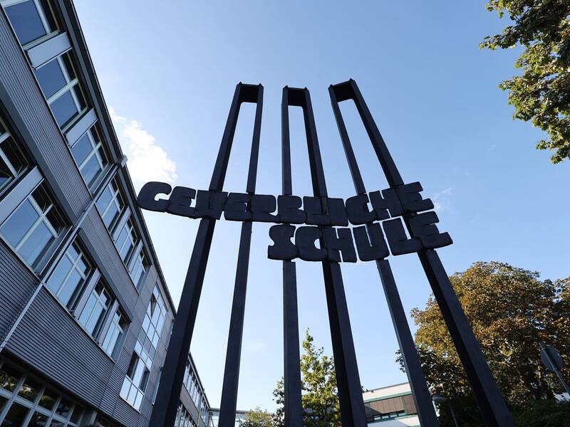 Gewerbliche Schule. Ein Schild mit der Aufschrift "GEWERBLICHE SCHULE" steht zwischen hohen Stahlträgern vor einem großen Gebäude unter blauem Himmel.