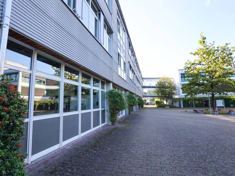 Gewerbliche Schule. Ein moderner Schulhof mit einem großen, gepflasterten Bereich und verschiedenen Schulgebäuden sowie Bäumen im Hintergrund.