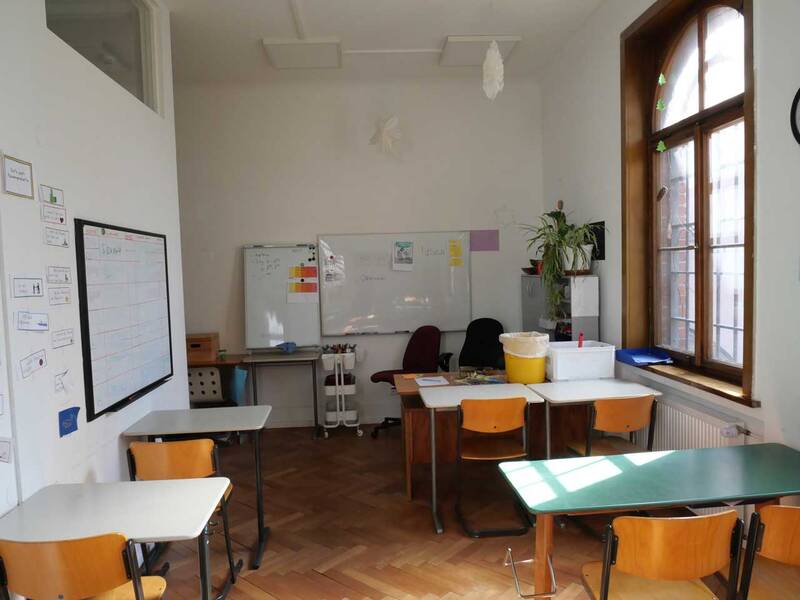 Ein heller Klassenraum mit Holzboden, mehreren Tischen und Stühlen, einer Tafel und einem großen Fenster.