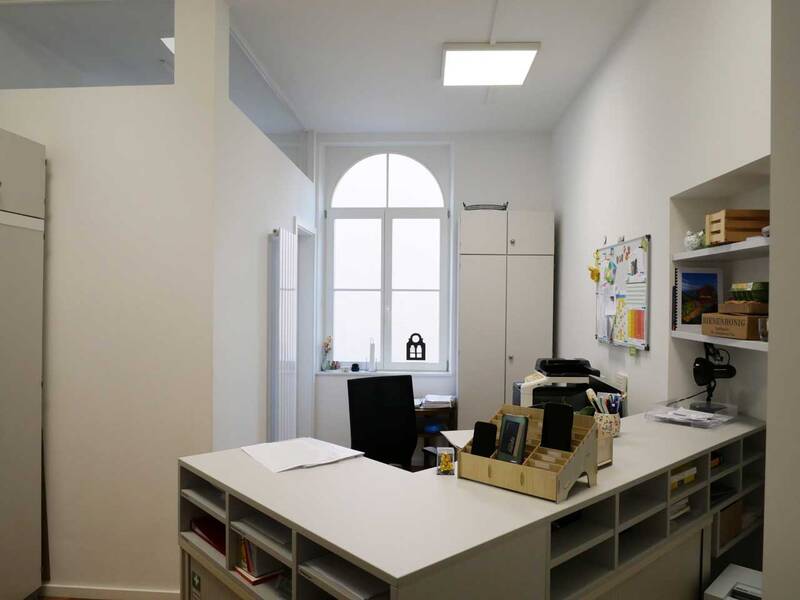 Das Bild zeigt ein kleines, ordentliches Büro mit einem Schreibtisch, einem Regal, einem Fenster im Hintergrund und einer Pinnwand an der rechten Wand.