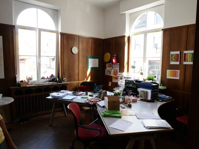Ein ordentliches Büro mit Holzwänden, auf dessen Schreibtisch viele Dokumente, Ordner und Pflanzen liegen, ist von großen Fenstern mit Rundbögen erleuchtet.