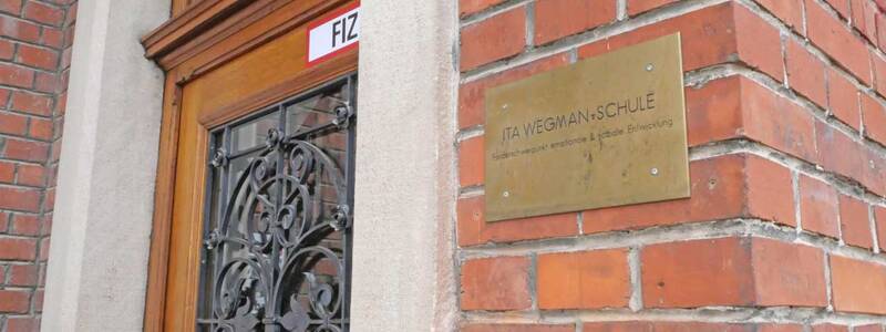 Eine Ziegelwand mit einem Messingschild, das "Ita Wegman Schule" zeigt, neben einer Holztür mit kunstvollem schwarzen Metallgitter.