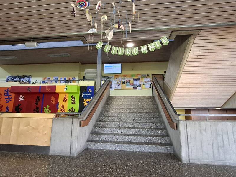 Das Bild zeigt das farbenfrohe Foyer eines Schulgebäudes mit einer zentralen Treppe und einem „Willkommen“-Banner.