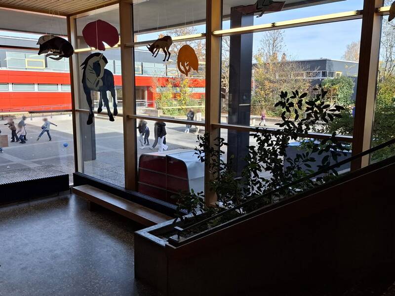 Das Bild zeigt einen Blick aus dem Schulgebäude auf den Pausenhof, wo Kinder spielen und Schüler vorbeigehen.