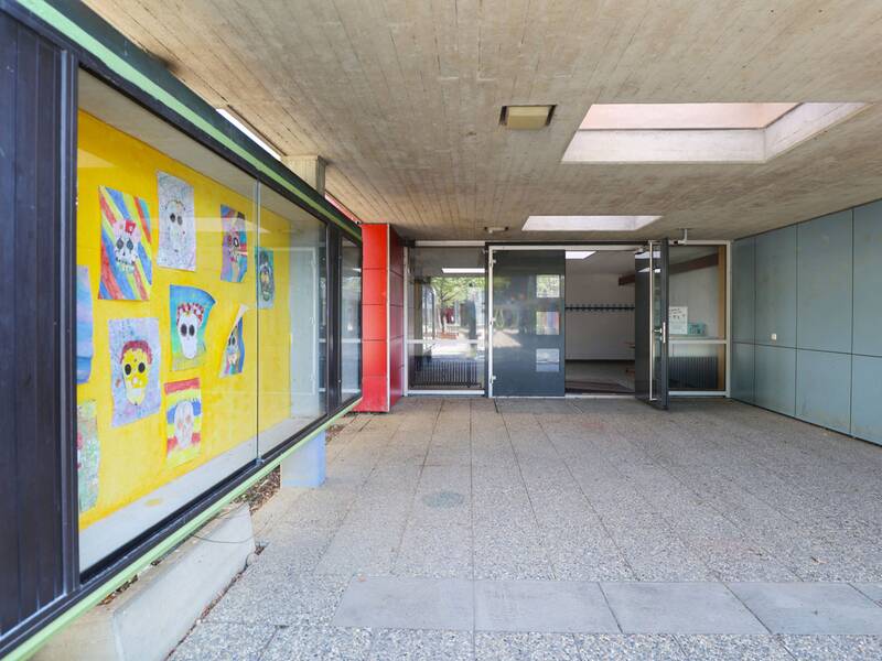 Neugreuthschule. Ein überdachter Eingangsbereich mit Glastüren und einer Wand mit bunten Kinderzeichnungen.