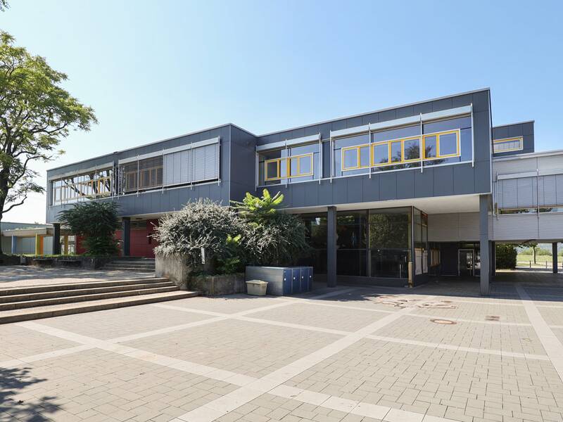 Neugreuthschule. Das Bild zeigt ein modernes zweistöckiges Gebäude mit grauer Fassade, gelben Fenstern und einer breiten Treppe, umgeben von Bäumen und Pflanzkübeln.