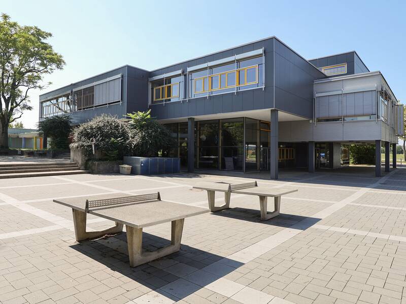 Neugreuthschule. Ein modernes, graues Gebäude mit großen Fenstern, umgeben von Bäumen und Pflanzen, steht auf einem gepflasterten Platz, der zwei Tischtennisplatten beinhaltet.