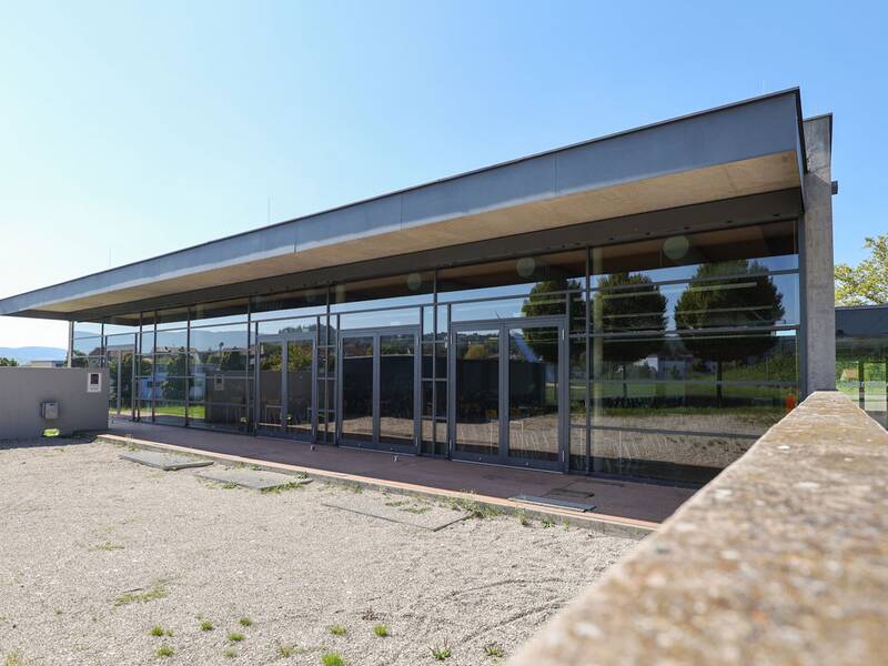Neugreuthschule. Die Mensa. Ein modernes, eingeschossiges Gebäude mit großen Glasfronten steht in einer Umgebung mit Kiesboden und vereinzeltem Graswuchs.