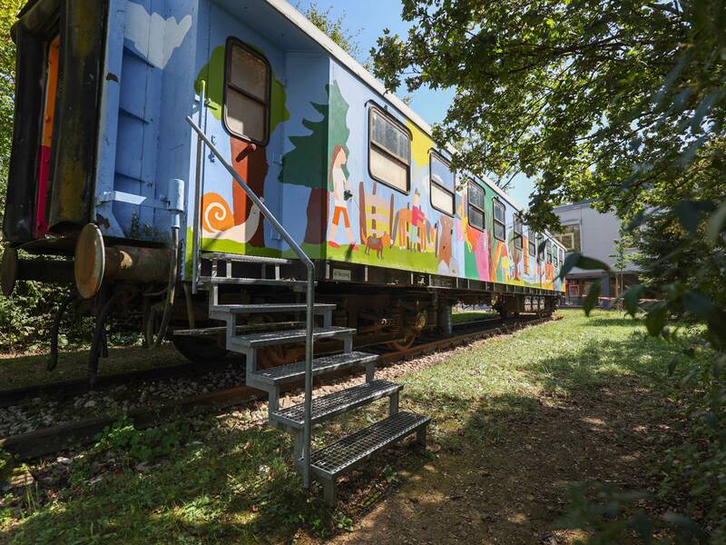 Neugreuthschule. Ein bunter, künstlerisch bemalter Eisenbahnwaggon steht auf einem Gleis inmitten von grüner Vegetation, mit einer Metalltreppe, die zum Eingang führt.