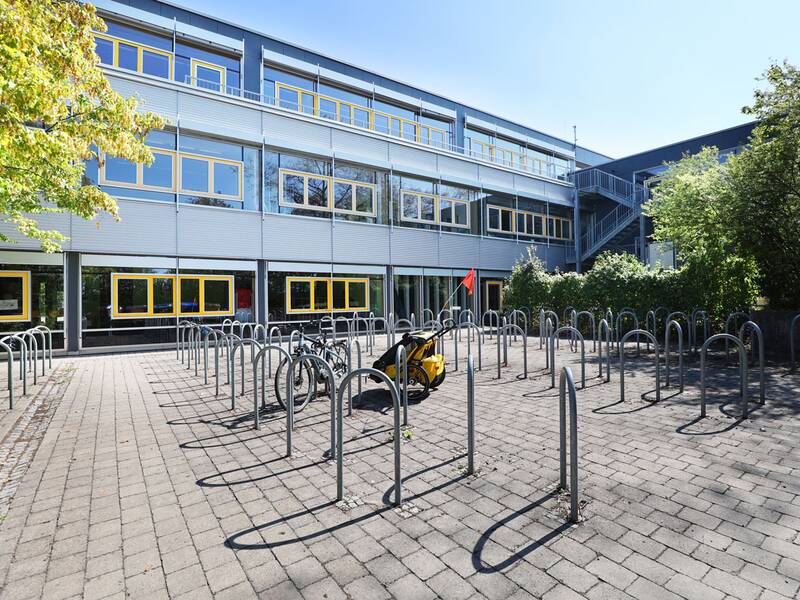 Neugreuthschule. Vor einem modernen Gebäude mit großen Fenstern stehen zahlreiche Fahrradständer auf einem gepflasterten Platz, in denen einige Fahrräder geparkt sind.