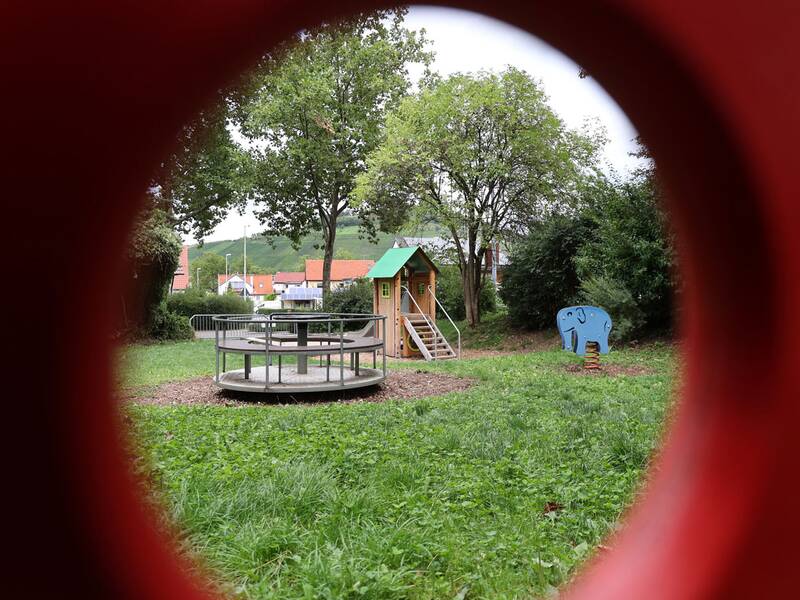 Blick durch ein rundes rotes Objekt auf ein Karussell, ein Spielhaus mit grüner Dachspitze und ein Wipptier in Elefantenform.