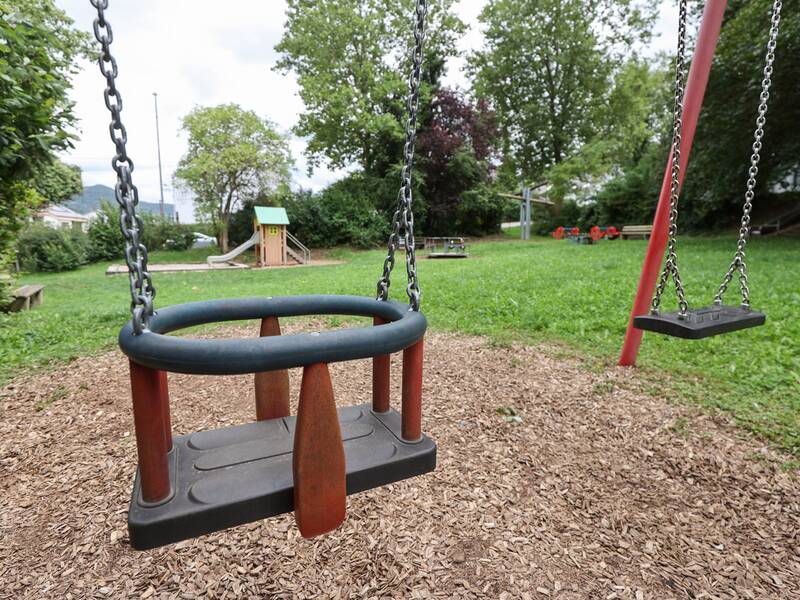Zwei Schaukeln, eine davon mit Kleinkindersitz, hängen an einem roten Gestell, im Hintergrund sind weitere Spielgeräte und Bäume zu sehen.