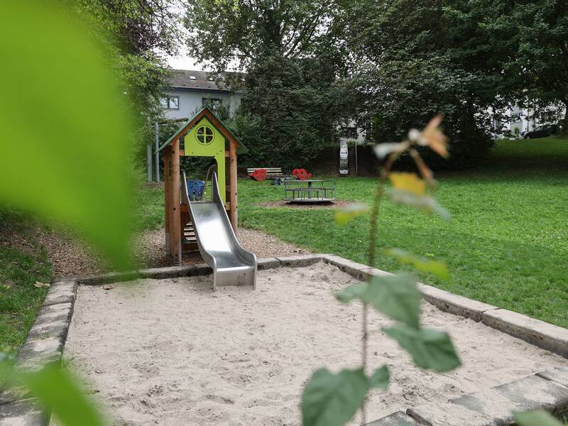 Im Vordergrund ein Sandkasten mit Rutsche, im Hintergrund Wippe, Karussell und eine weitere Rutsche, umgeben von Wiese und Bäumen.