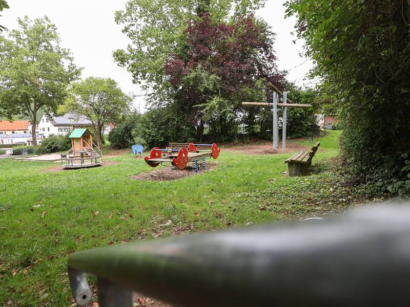 Ein Spielplatz mit Rasenfläche, im Vordergrund eine rote Wippe mit vier Sitzen auf Federn, im Hintergrund ein kleines Spielhaus und zwei Schaukeln. Bäume und Büsche umgeben den Platz.