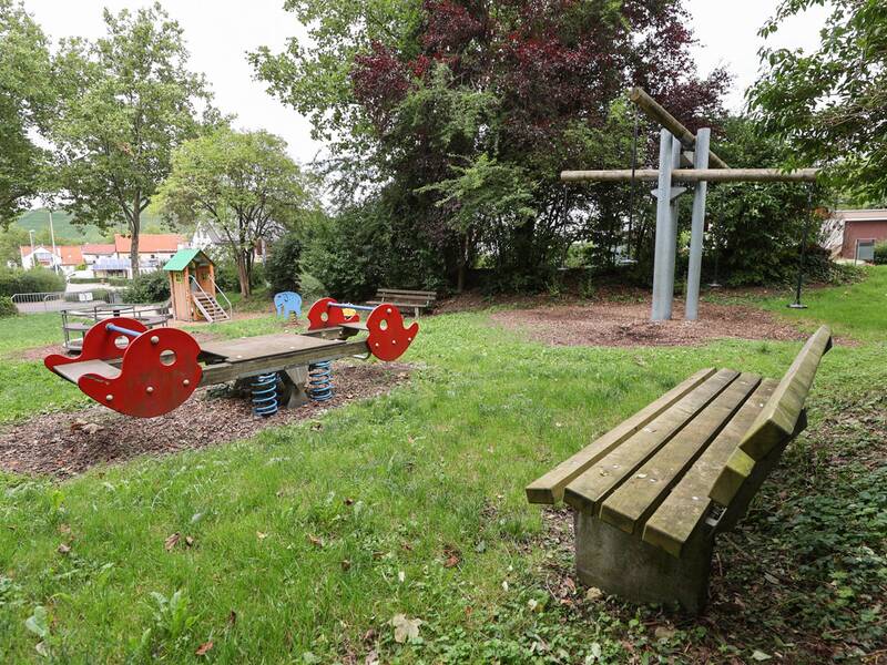 Die Wippe, das Spielhaus und die Doppelschaukel sind zentral zu sehen, vorne steht eine Holzbank.
