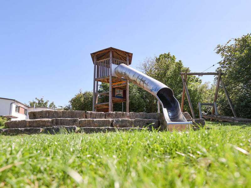 Der hölzerne Spielturm mit überdachter Plattform und langer Röhrenrutsche aus Metall, davor Naturstein-Stufen.