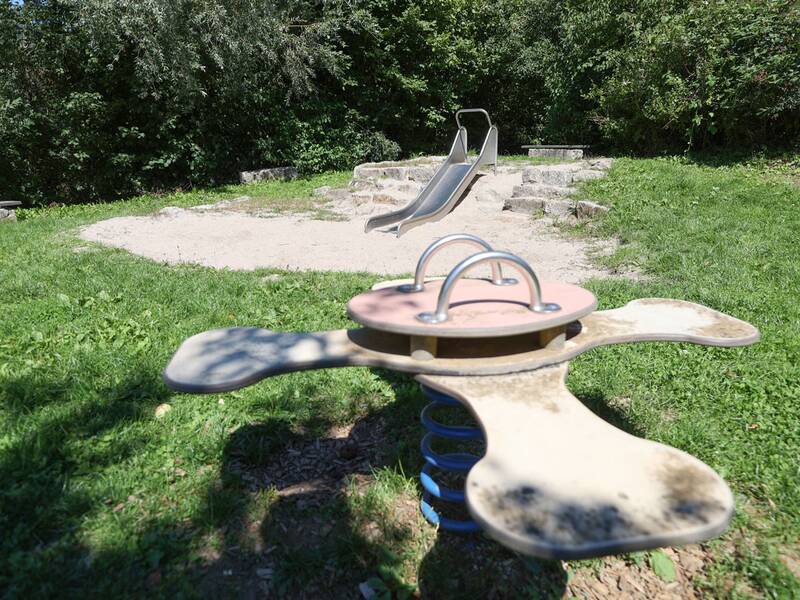 Eine kleine Rutsche aus Metall, eingebettet in eine Sandfläche mit Naturstein-Stufen, im Vordergrund ein Wipp-Tier mit vier Armen.