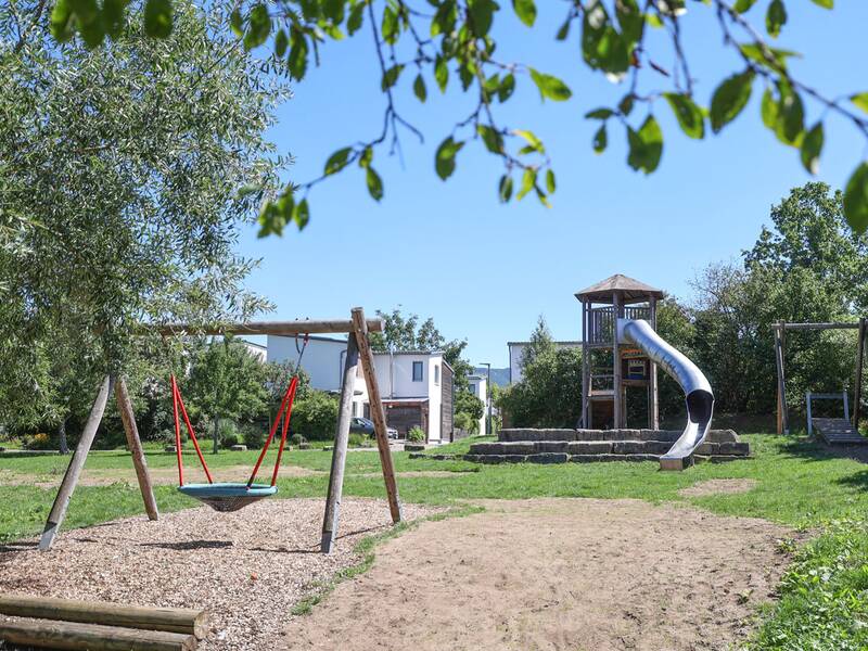 Ein großer Spielplatz mit Nestschaukel, langer Röhrenrutsche und Seilbahn, eingerahmt von viel Grün