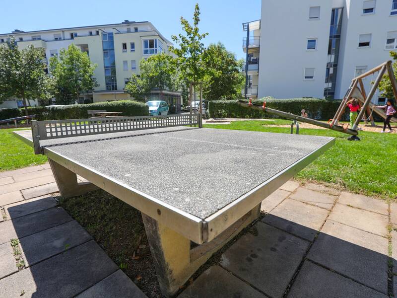 Eine Tischtennisplatte aus Beton steht auf einem gepflasterten Bereich, im Hintergrund spielende Kinder.