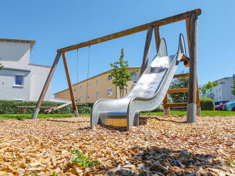 Eine Rutsche aus Metall ist in ein Holzgerüst mit Schaukel integriert, umgeben von Rindenmulch und Wohnhäusern.