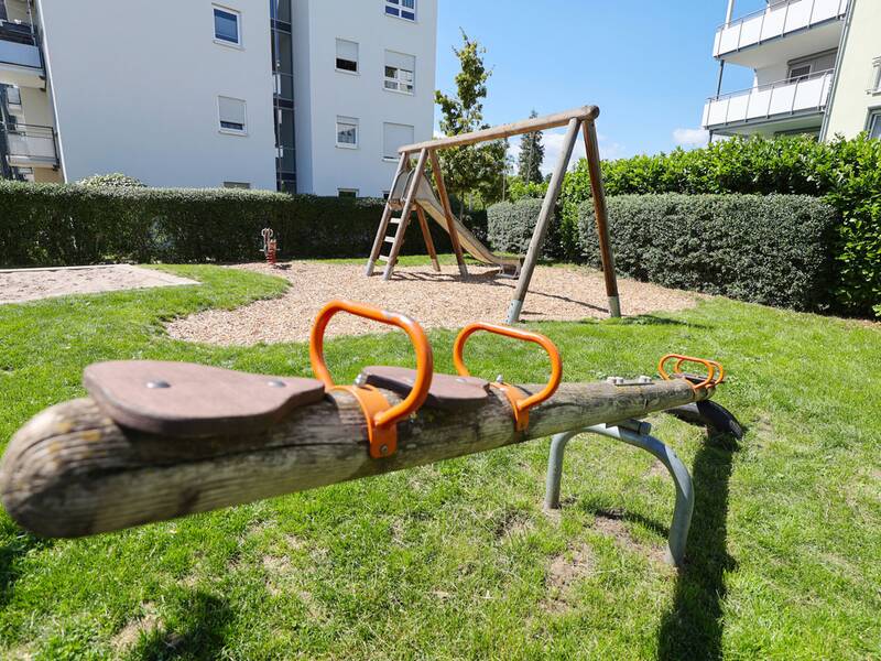 Blick auf einen Spielplatz mit Rutsche und Schaukel, im Vordergrund eine Wippe aus Holz