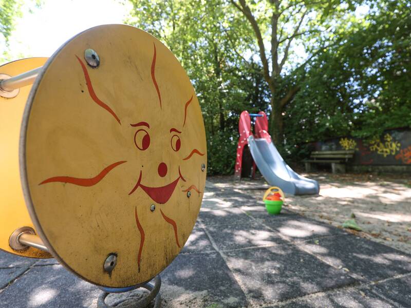 Ein gelbes Spielgerät mit aufgemaltem Sonnengesicht steht auf Gummiplatten, im Hintergrund eine kleine Rutsche im Sand.