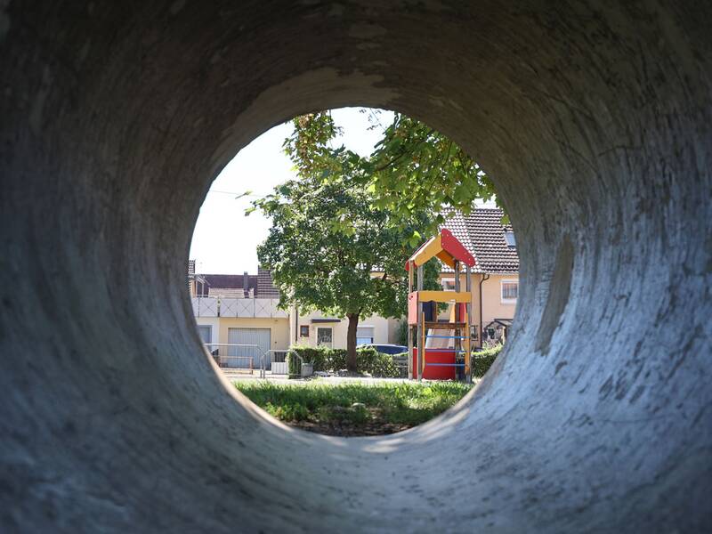 Blick durch ein großes Betonrohr auf den Spielplatz mit Kletterturm und Baum.