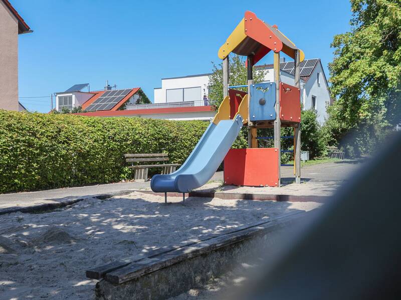 Der Kletterturm mit Rutsche steht direkt an einem Sandkasten, im Hintergrund Bänke und Häuser.