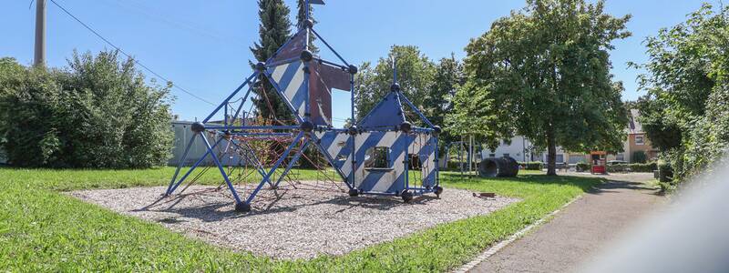 Weitwinkelaufnahme des Spielplatzes mit Klettergerüst, Schaukeln, Rutsche und Bäumen.