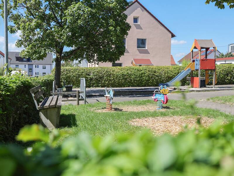 Spielplatz mit buntem Kletterturm, zwei Wipptieren, Bänken und Bäumen.