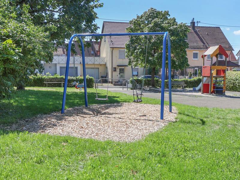 Zwei Schaukeln – eine normale, eine mit Babysitz – vor dem Kletterturm, umgeben von Häusern.