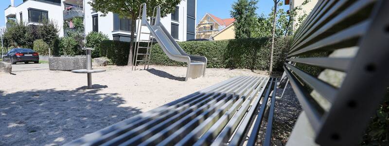 Eine Metallrutsche steht auf einem Sandplatz zwischen Bäumen, im Vordergrund eine Metallbank, im Hintergrund moderne Wohnhäuser.