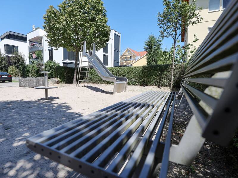 Eine Metallrutsche steht auf einem Sandplatz zwischen Bäumen, im Vordergrund eine Metallbank, im Hintergrund moderne Wohnhäuser.