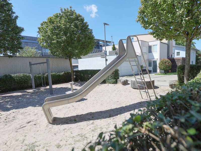 Seitlich aufgenommene Rutsche mit Leiter auf einem Sandspielplatz, dahinter ein Schaukelgestell und ein Baum.