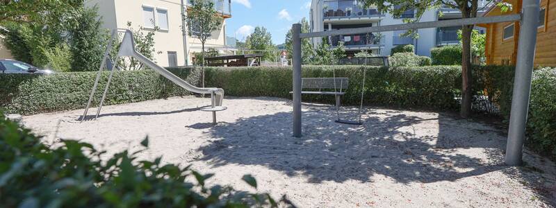 Spielplatz mit Rutsche, Schaukel und Sitzbank, alles auf Sandboden, umgeben von Hecken und Wohnhäusern.