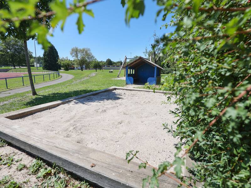 Ein Sandkasten mit Holzrahmen steht neben einem kleinen blauen Spielhäuschen.