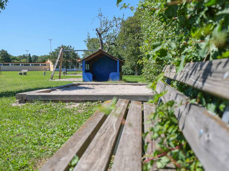 Ein Sandkasten und ein Häuschen sind von einer Holzbank und grüner Wiese umgeben.
