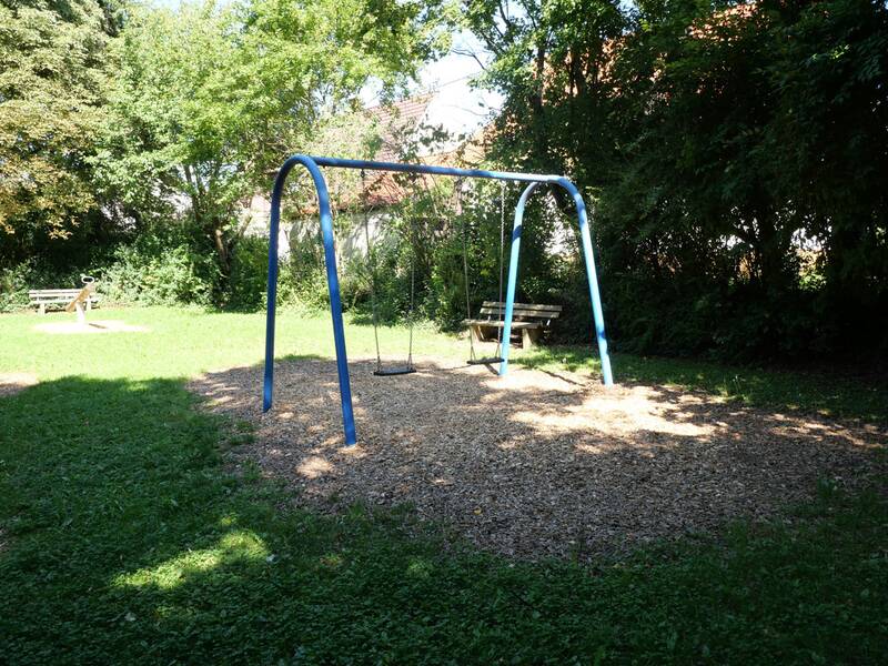 Eine leere Schaukel mit blauem Gestell steht auf einem mit Holzschnitzeln bedeckten Platz auf einem grünen Spielplatz, umgeben von Bäumen und einer Bank im Hintergrund.