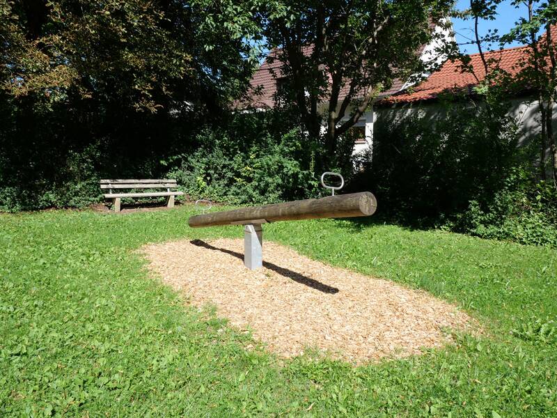 Ein Kinderspielplatz mit einer Wippe auf einer Mulchfläche, umgeben von grüner Wiese und Bäumen, mit einer Bank im Hintergrund.
